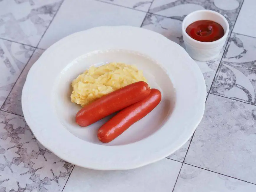 A plate of mashed potatoes and sausages with a small cup of ketchup on a decorative tile table.