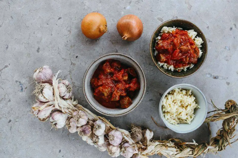 Top view of a rustic meal with garlic, onions, and bowls of rice and stew on a stone surface.