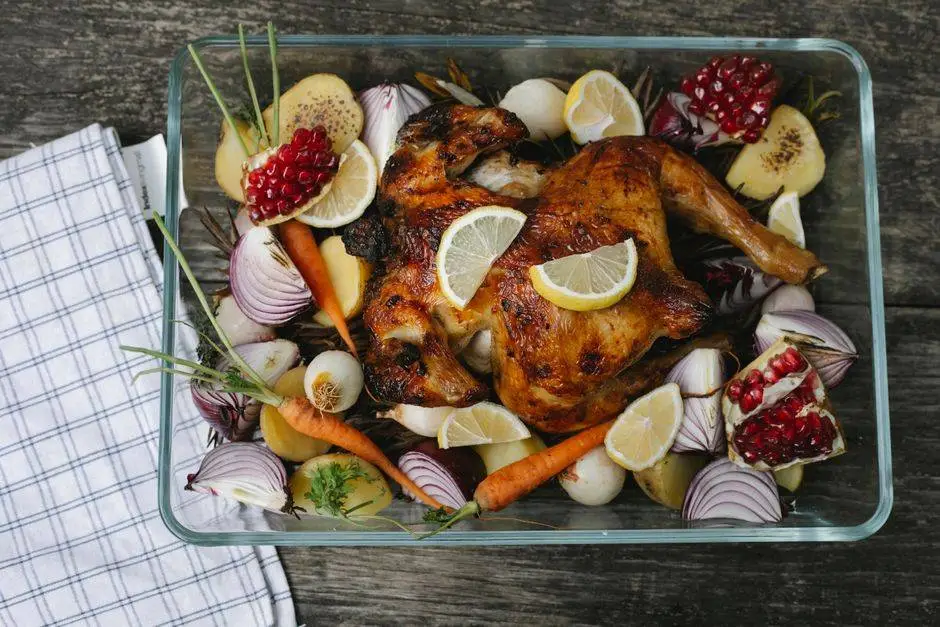 Top view of appetizing baked chicken with onion and lemon slices near fresh pomegranate pieces and whole carrots in baking dish